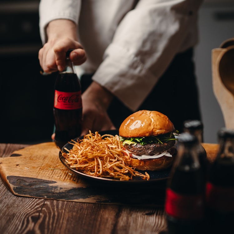Burger me patate të skuqura në pjatë dhe dorë që mban shishe Coca-Cola.