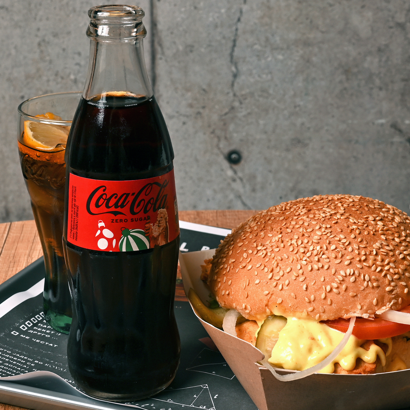 Shishe Coca-Cola Zero me burger dhe gotë me limon në një tavolinë druri.