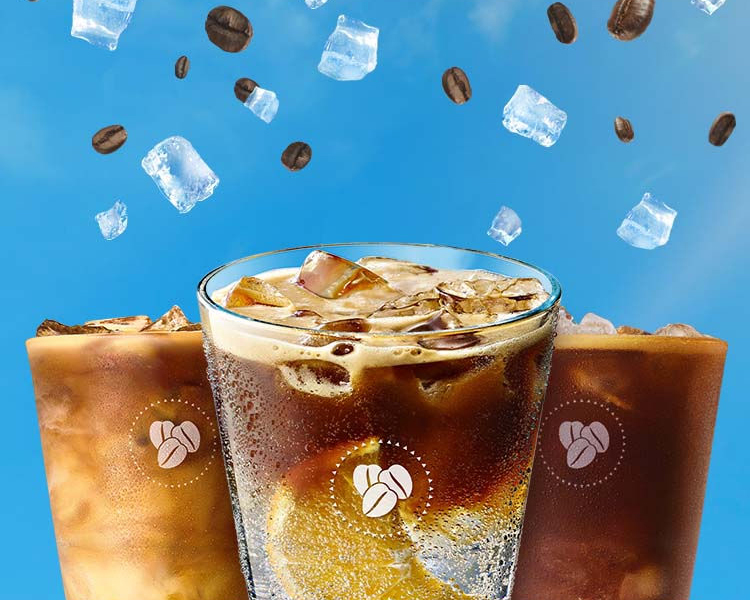 Tres vasos de cristal con diferentes tipos de cafés fríos sobre un cielo celeste con lluvia de hielos y granos de café.
