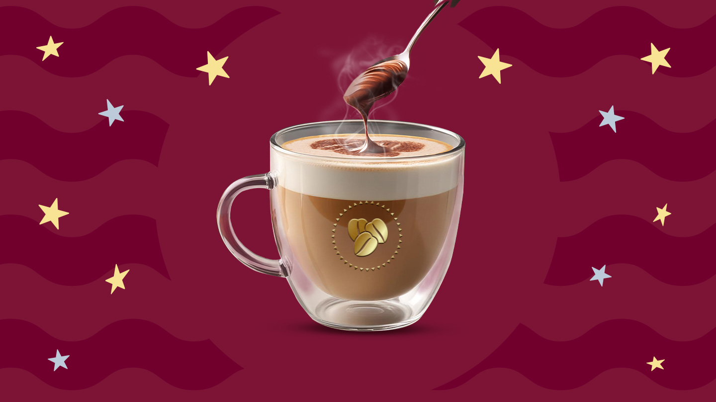 Una taza de cristal con café latte caliente y una cuchara por encima agregando crema de avellanas sobre un fondo en tonos guinda.