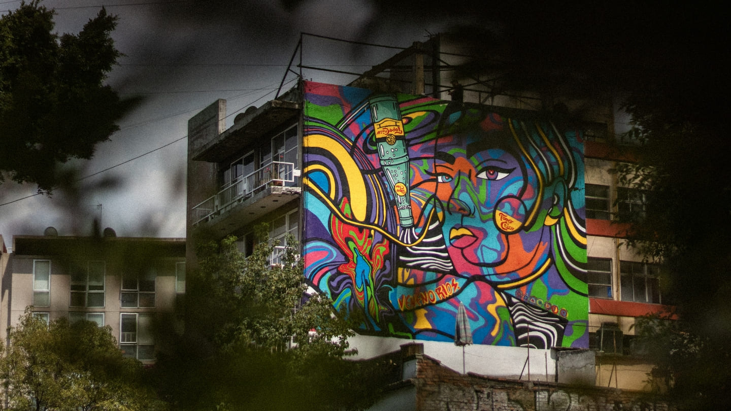 Un edificio con un mural colorido y abstracto de un rostro se alza contra un cielo nocturno. Los árboles en primer plano enmarcan la escena.