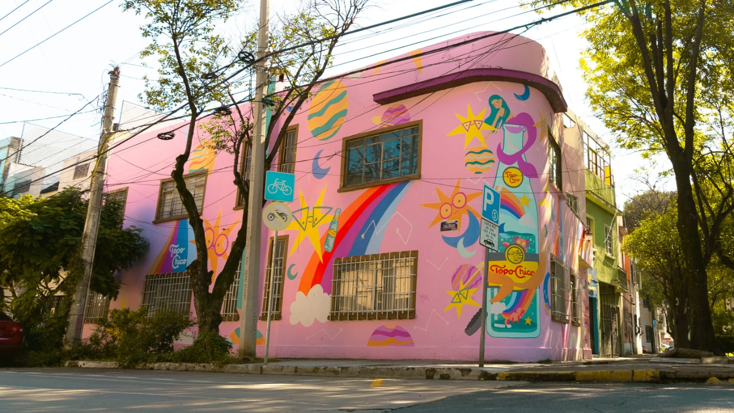 Un edificio rosa con murales coloridos, incluyendo un arcoíris y diseños celestiales, se encuentra en una calle de la ciudad. Árboles y cables de electricidad son visibles en primer plano.
