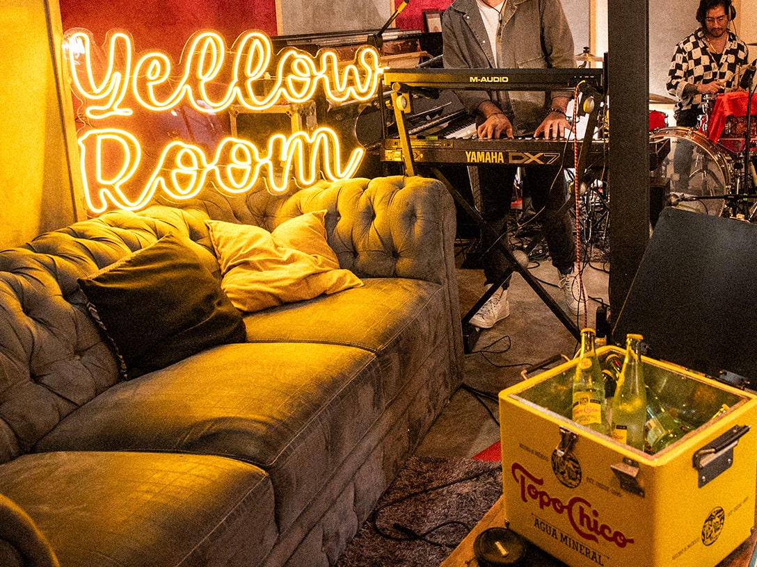 Estudio de música en el que hay un sillón color amarillo, una heladera con botellas de vidrio de Topo Chico, al fondo hay un baterista y un tecladista y al lado hay un cartel luminoso que dice: Yellow room.