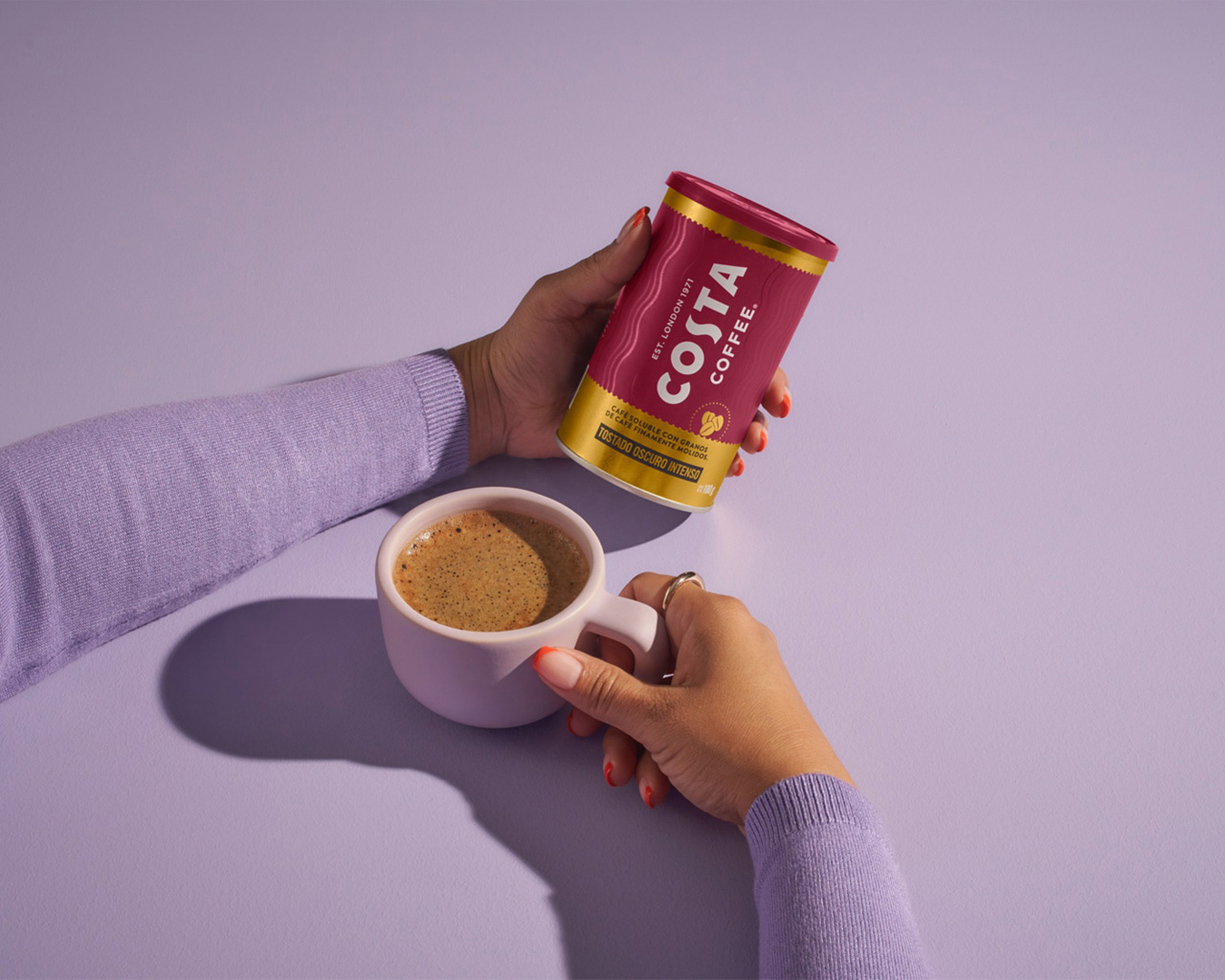 Una persona agarrando una taza de café y con la otra mano sosteniendo una lata de café soluble Costa Coffee.