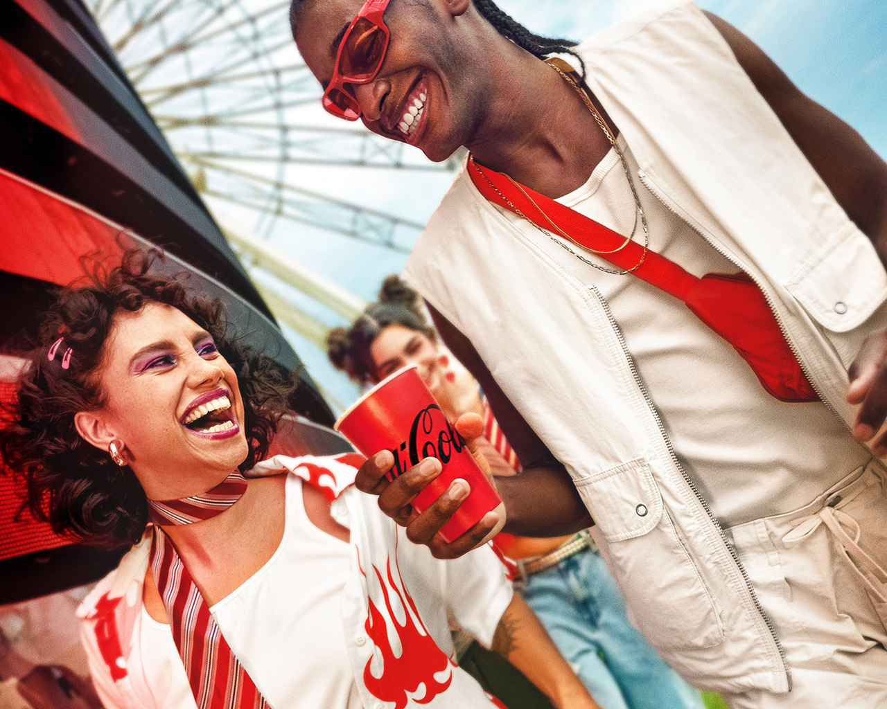 Dos personas disfrutando de una Coca-Cola, sonriendo y riendo. Una persona tiene el pelo rizado con accesorios rosados y lleva una bufanda a rayas, mientras que la otra persona tiene trenzas, usa gafas de sol, un chaleco blanco y una bolsa cruzada roja. Líneas gráficas rojas mejoran el ambiente festivo.