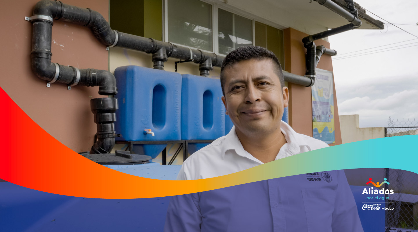 Hombre sonriente frente a sistema de recolección de agua de lluvia en escuela, parte de la iniciativa 'Aliados por el Agua' de Coca-Cola México.