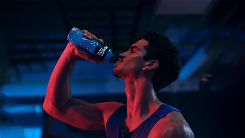 Sportist pi një pije Powerade blu në palestër me ndriçim blu dhe të kuq.