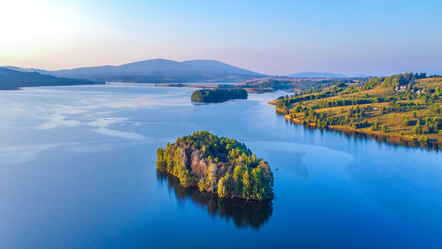 Pogled iz vazduha na mirno jezero sa malim zelenim ostrvom u prvom planu, okruženo šumovitim obalama i planinama u daljini