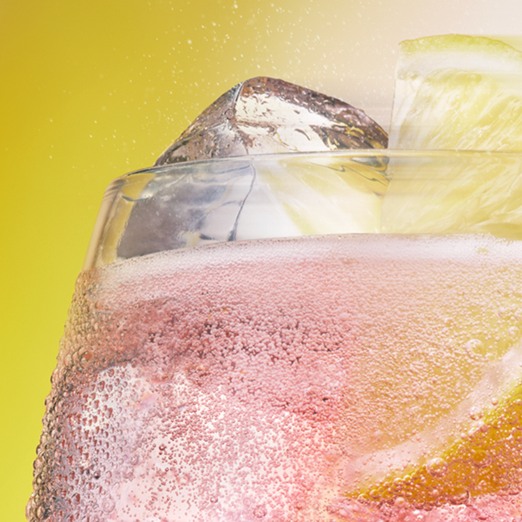 Primer plano de una copa efervescente de rhubarb gin tonic con cubos de hielo y una rodaja de limón sobre un fondo amarillo.