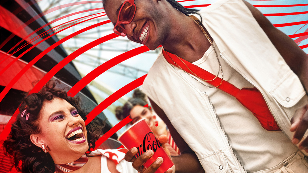 Dos personas riendo mientras disfrutan de Coca-Cola en un evento al aire libre, con decoraciones rojas en el fondo.